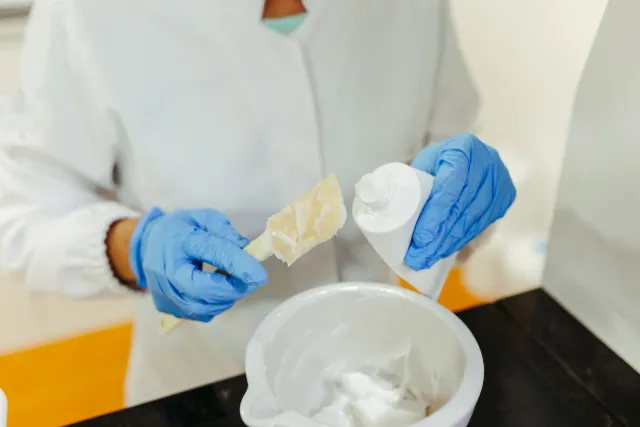 Gloved hands transferring a white skincare cream into a mixing bowl in a laboratory.