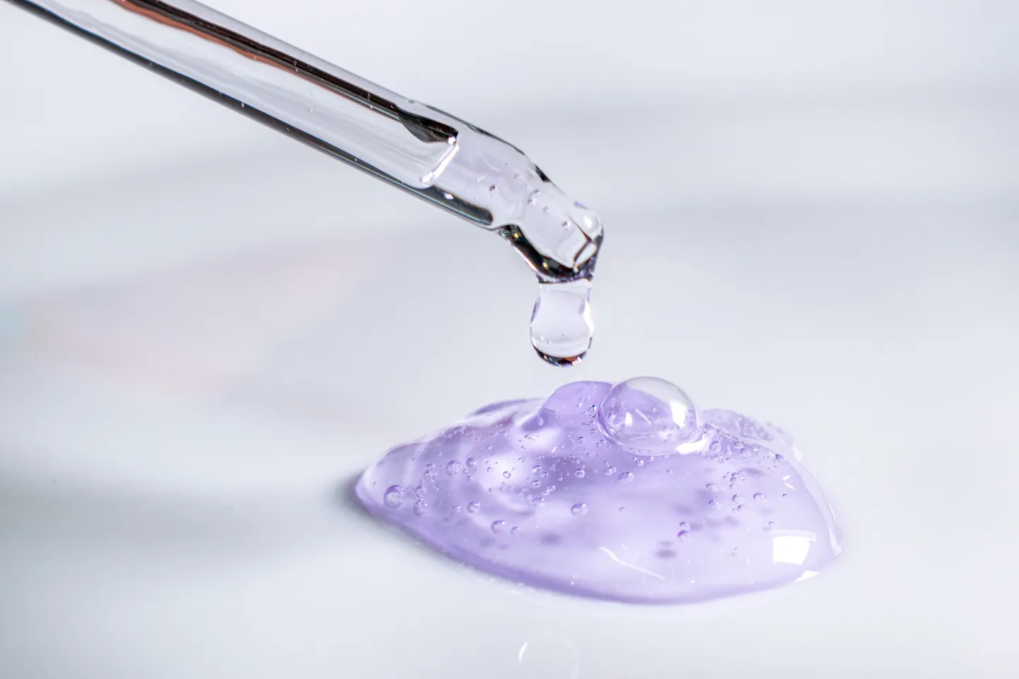 A glass pipette releasing a drop of clear lilac gel onto a white surface.