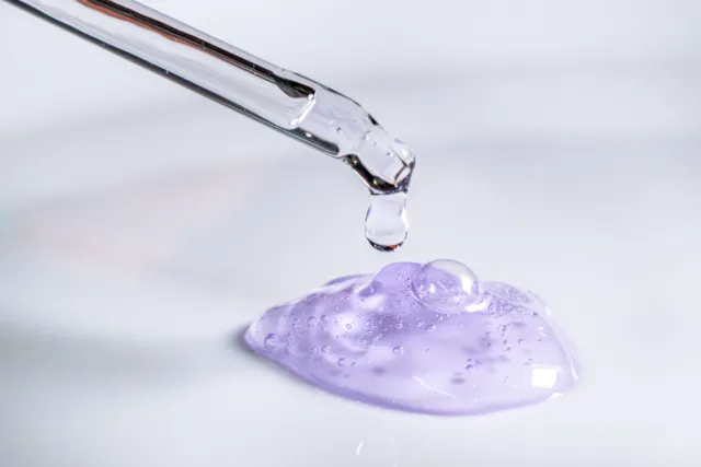 A glass pipette releasing a drop of clear lilac gel onto a white surface.