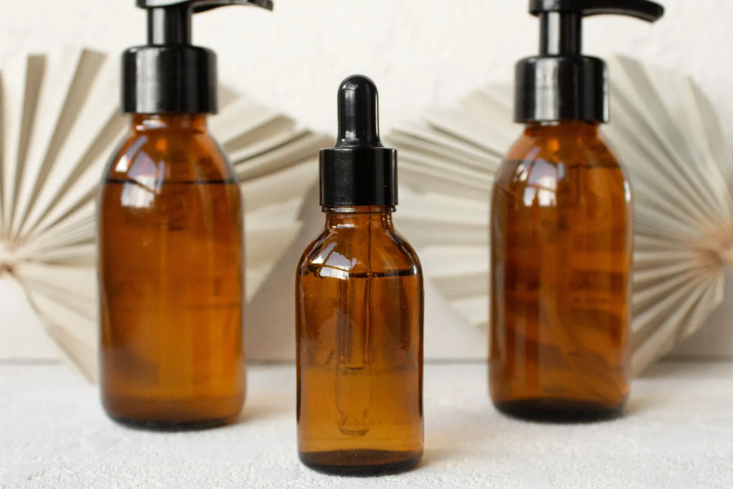 Three amber glass bottles, two with black pumps and one with a dropper, in front of a pale wall.